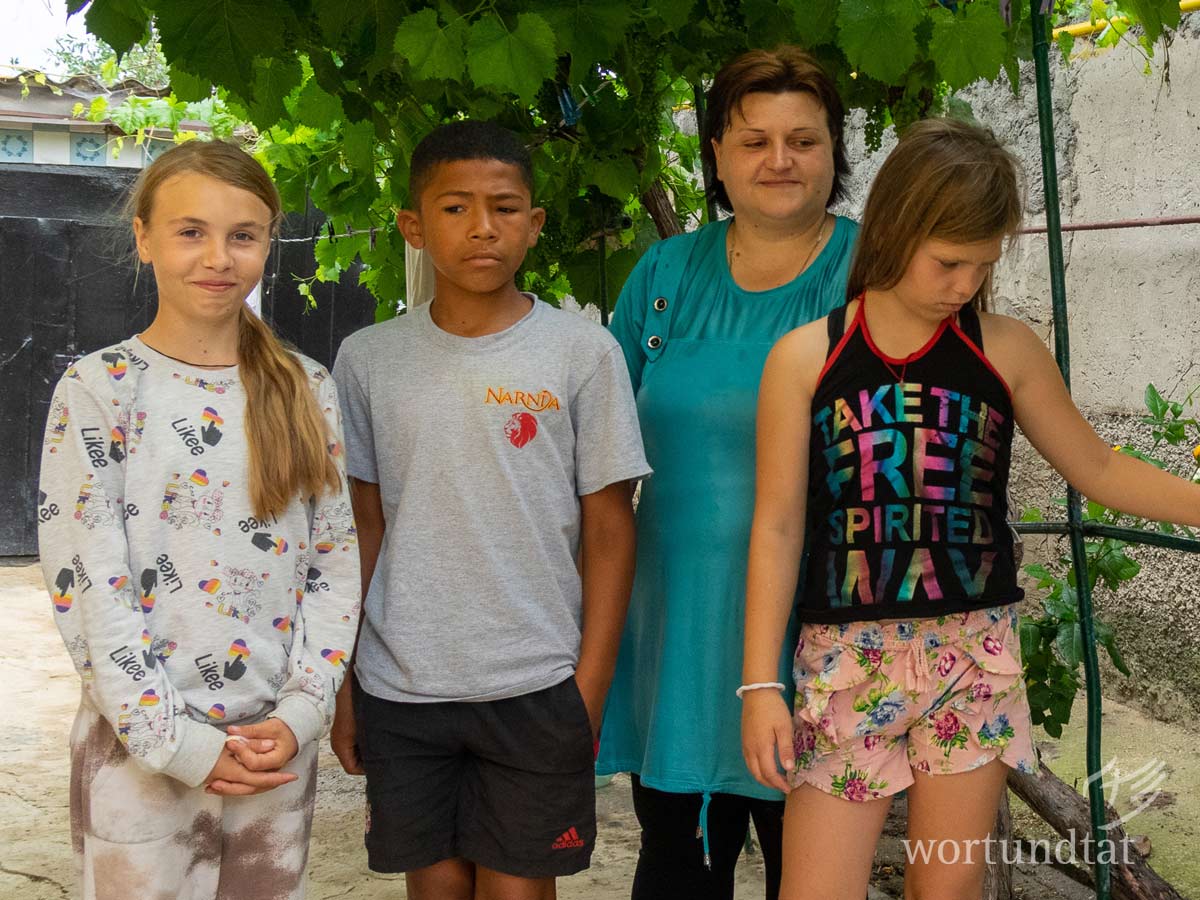 Sjuscha (12 Jahre), Oliver (11 Jahre), Valeria (11 Jahre) leben zusammen mit der Mutter der beiden Mädchen, einer Oma und einer weiteren Verwandten in einem Appartement mit etwa 30 Quadratmetern. Die Unterkunft ist in drei Zimmer und eine Küche aufgeteilt. Ein Holzofen heizt die Wohnung. Im Winter 2021/22 reichte das Holz nur zum einmaligen Heizen pro Tag. So kühlte das Haus bis zum Abend auf 10 Grad ab, bevor wieder angefeuert wurde. three children with mother